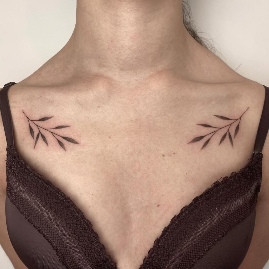 A simple and elegant collarbone tattoo featuring symmetrical leafy branches in soft black shading.