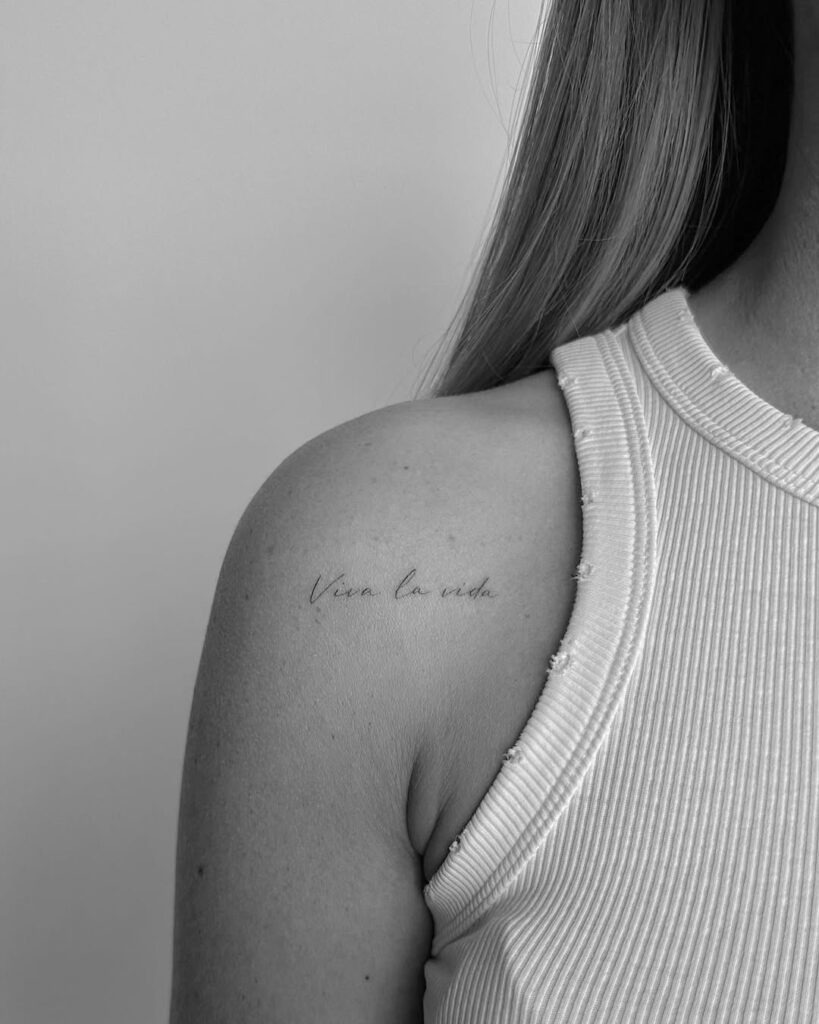 A simple yet impactful tattoo featuring the phrase "Viva la vida" in delicate cursive font, placed subtly near the collarbone.