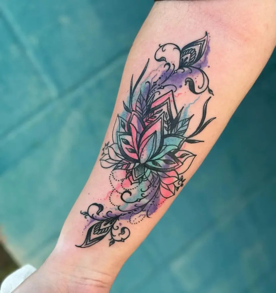 This tattoo showcases an intricate mandala pattern composed of floral elements, surrounded by a soft watercolor wash of purples and blues.