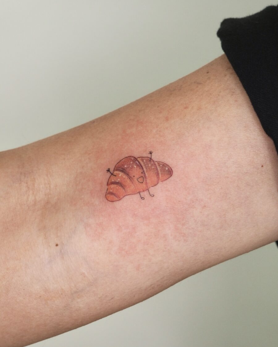 This tattoo showcases a cute, cartoonish croissant sitting playfully on the skin.