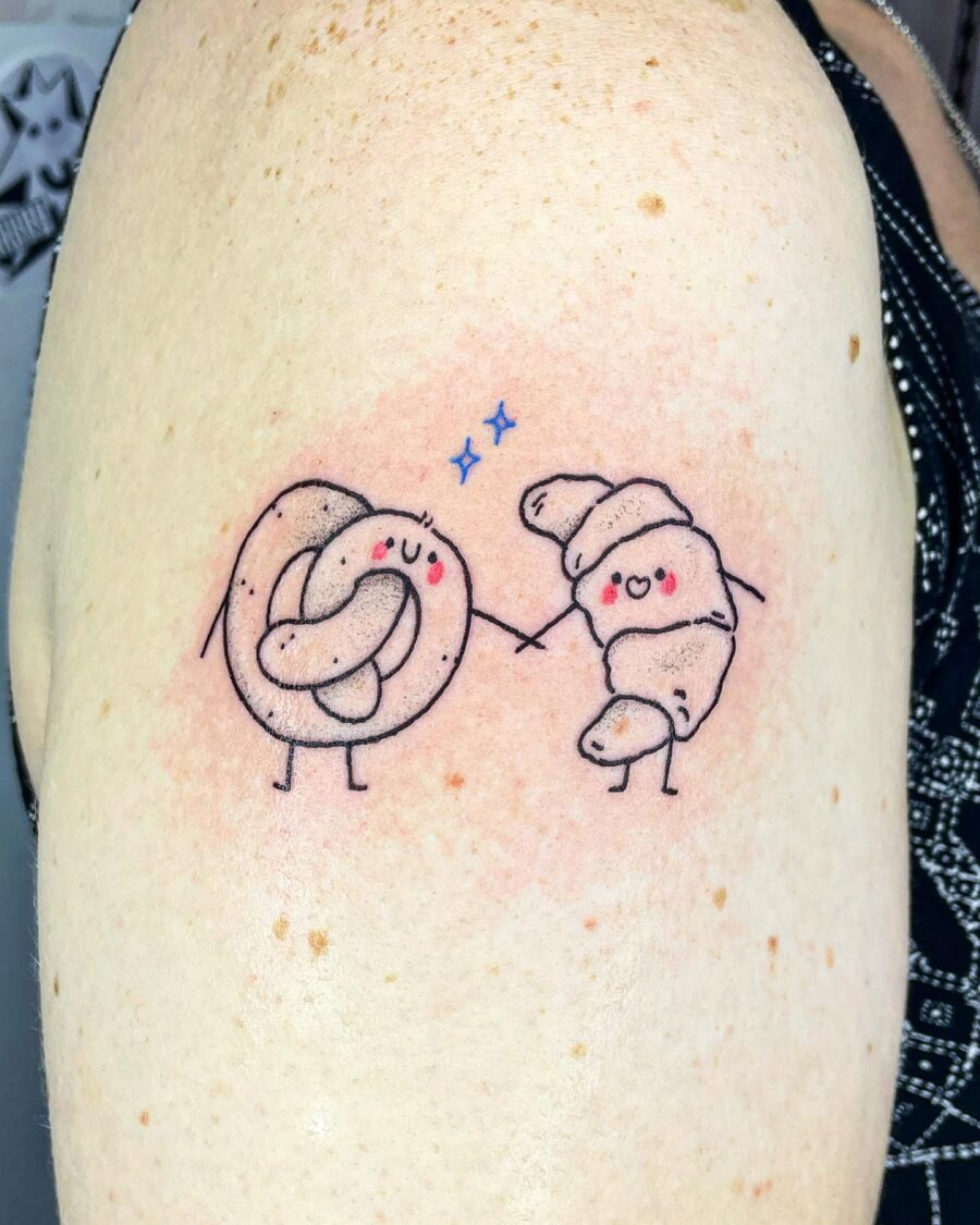 This tattoo features a cute croissant and a joyful pretzel holding hands, embodying a whimsical charm.