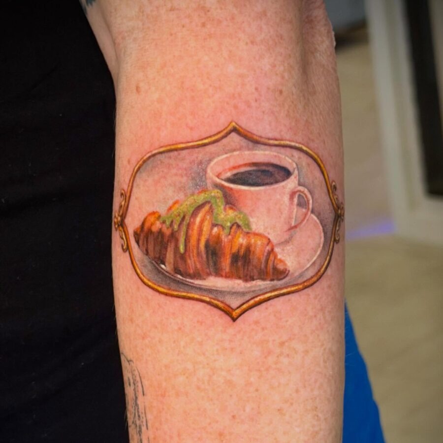 This tattoo features a beautifully detailed croissant resting on a plate next to a charming coffee cup.