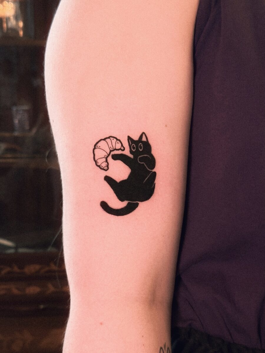 This tattoo features a playful black cat holding a croissant, showing a whimsical vibe.