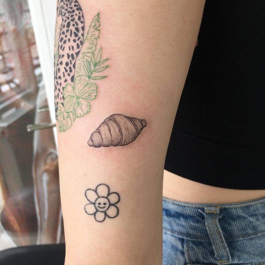This delightful tattoo showcases a croissant alongside a simple flower design.