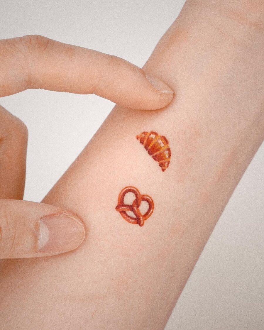 This image showcases two delightful tattoos on a hand: a croissant and a pretzel.