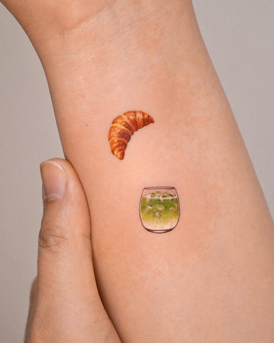 This tattoo features a delicious croissant and a whimsical glass of water, beautifully inked on the forearm.