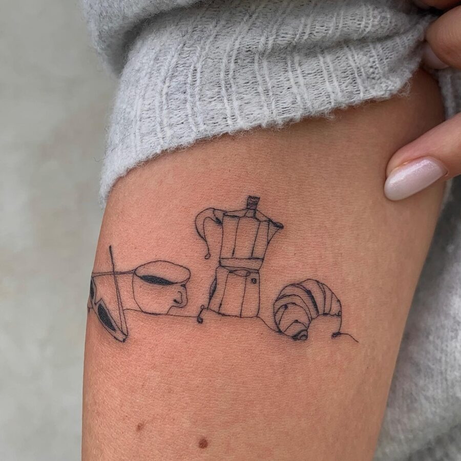 This tattoo features a whimsical design incorporating a croissant, a coffee pot, and some sunglasses.