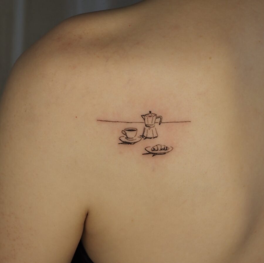 This tattoo features a minimalist design showing a coffee pot and cup alongside a croissant.