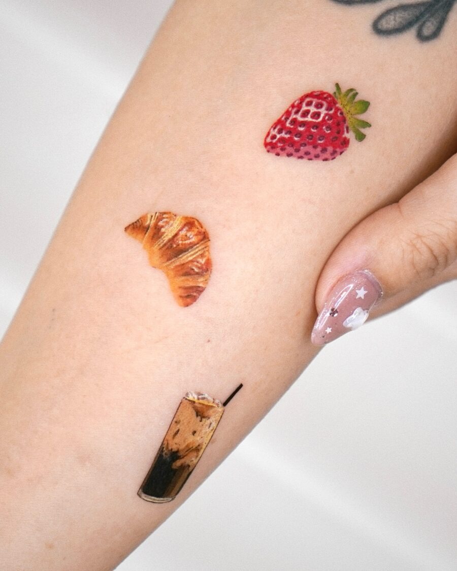 This delightful croissant tattoo is playfully surrounded by a juicy strawberry and a refreshing drink.