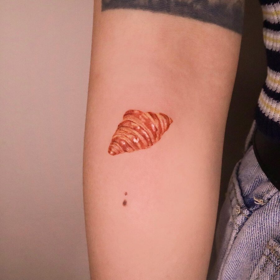 This tattoo features a beautifully realistic croissant, showing intricate details and warm tones.