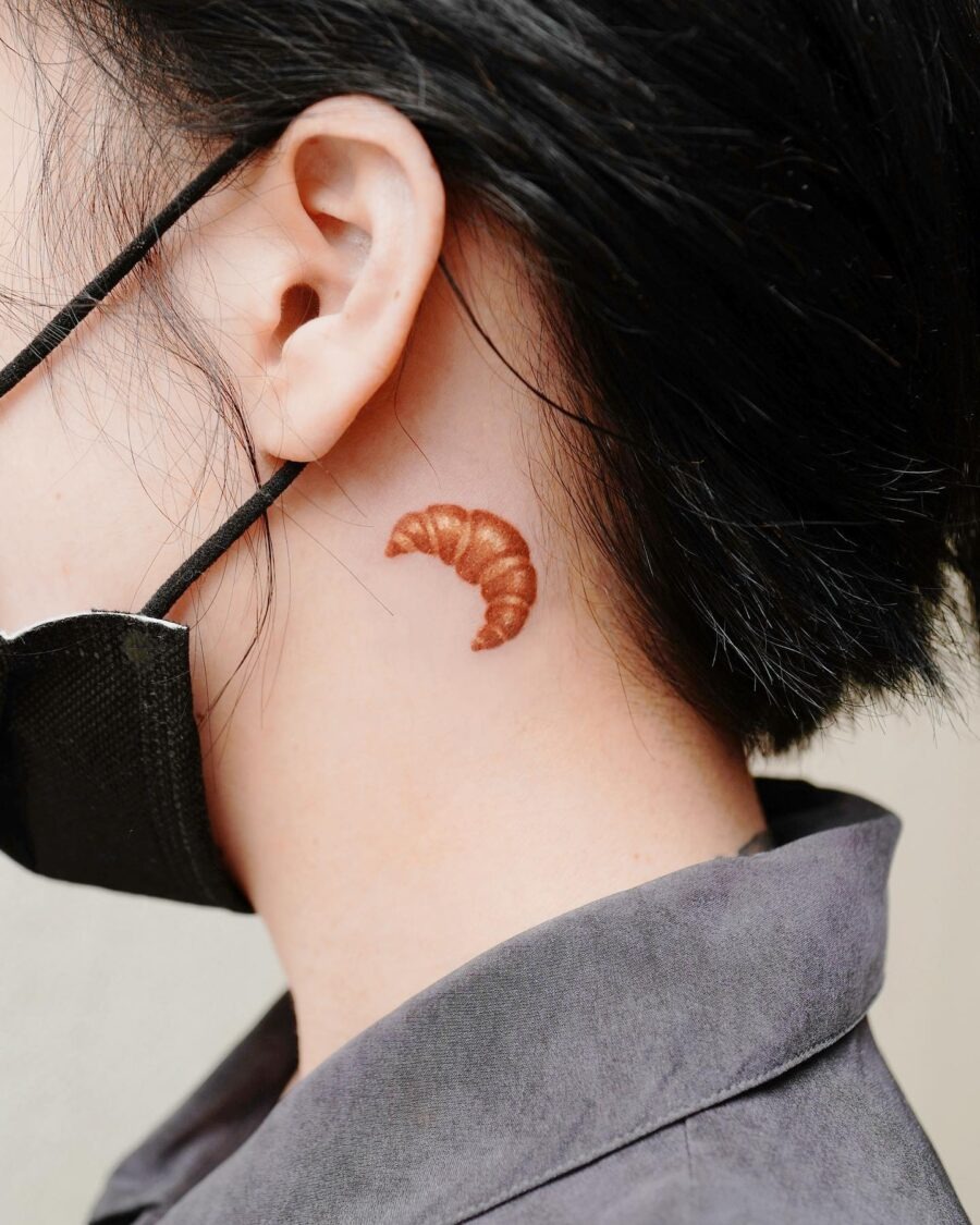 This artistic tattoo features a realistic croissant nestled at the nape of the neck.