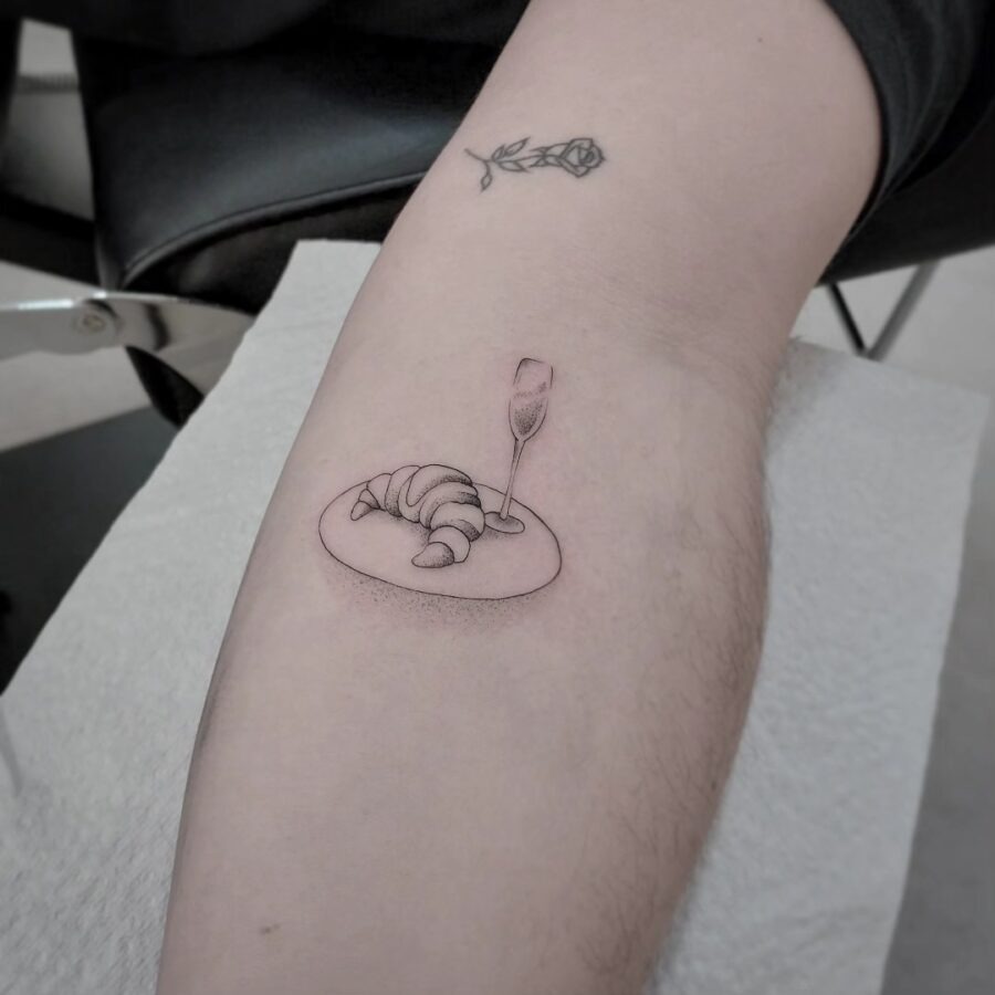 This cute tattoo features a croissant resting on a plate, elegantly paired with a champagne glass.