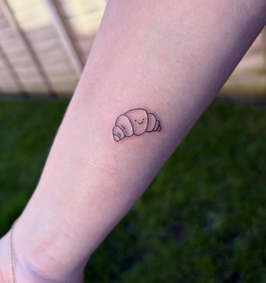 This cute croissant tattoo features a smiling face, adding a playful touch to the design.