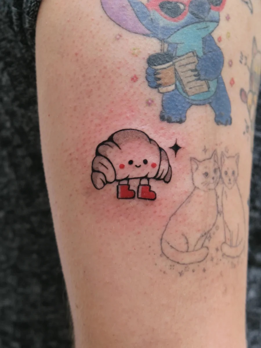 This tattoo features a cute croissant character, complete with whimsical red shoes and a joyful expression.