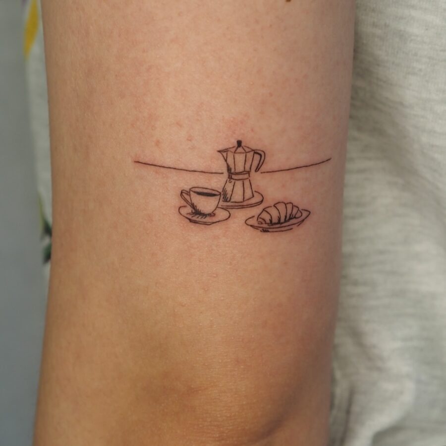 This tattoo features a minimalist design of a croissant alongside a classic moka pot and cup of coffee.