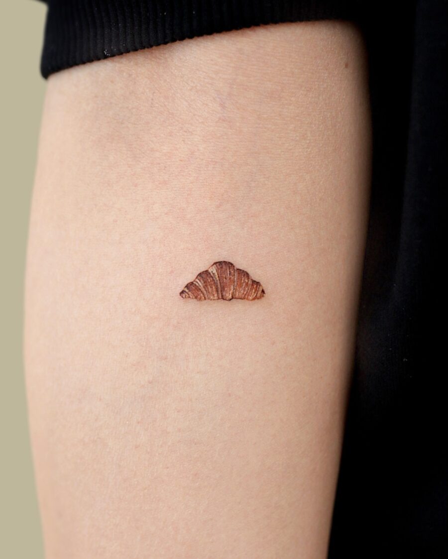 This charming croissant tattoo showcases a small, delicately designed croissant on the arm.