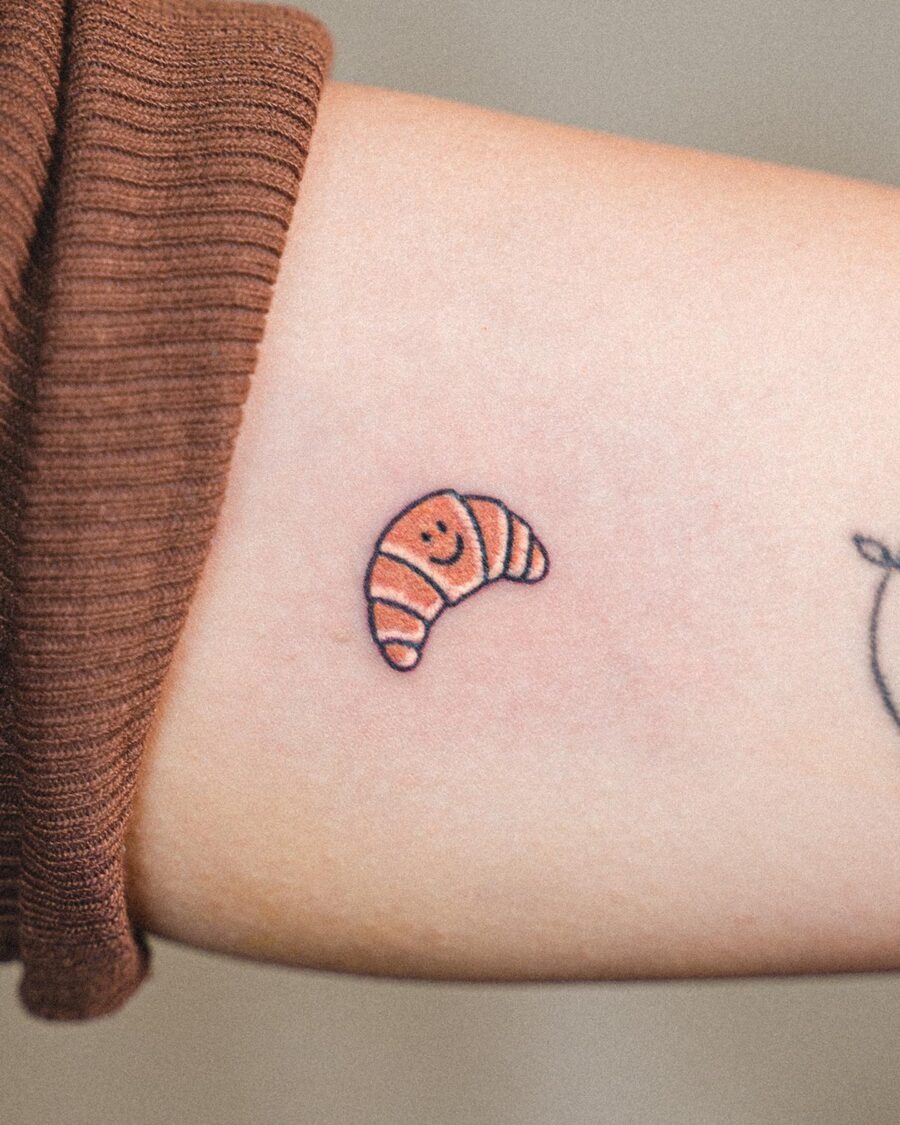 This delightful croissant tattoo features a cute smiling face, bringing a friendly vibe to the design.