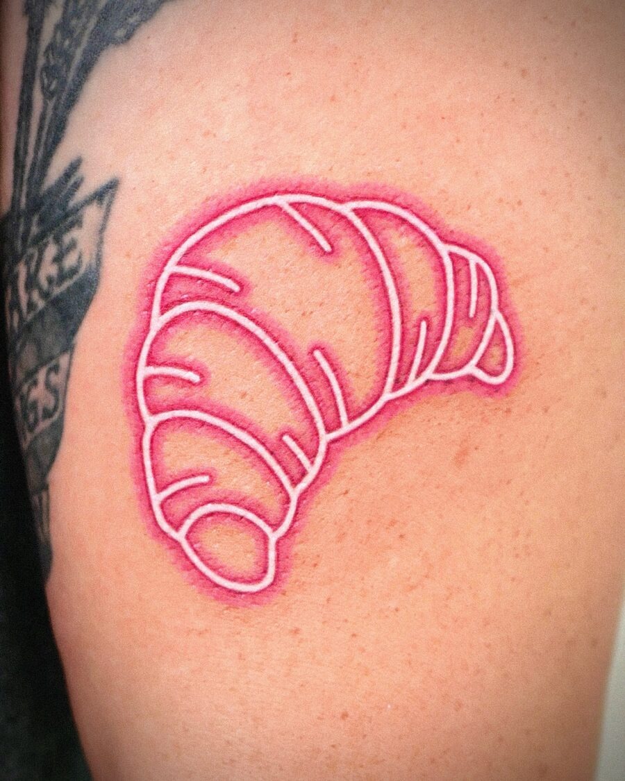 This tattoo features a sleek, minimalistic outline of a croissant in a pink hue.