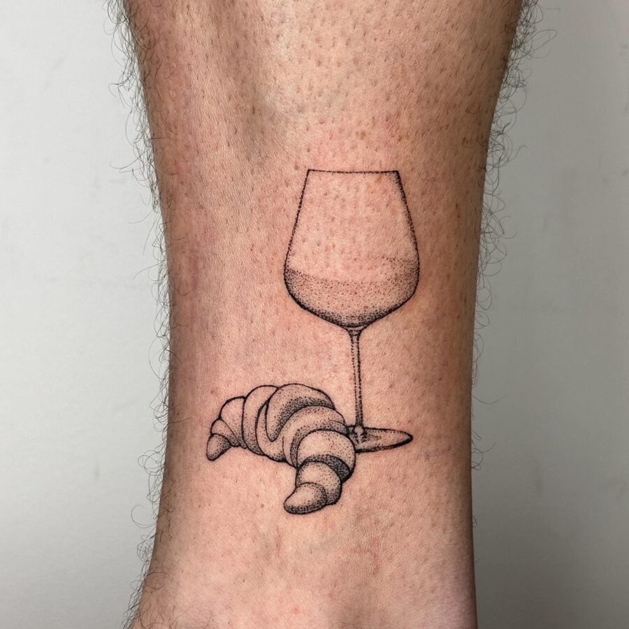 This tattoo features a delicate croissant nestled beside a wine glass, blending culinary delight with artistry.