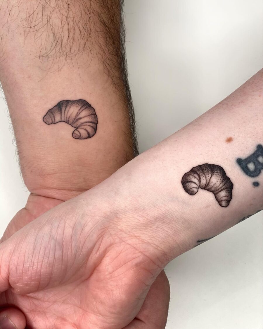 This image showcases two stylish croissant tattoos, elegantly inked in black on the forearms of both individuals.