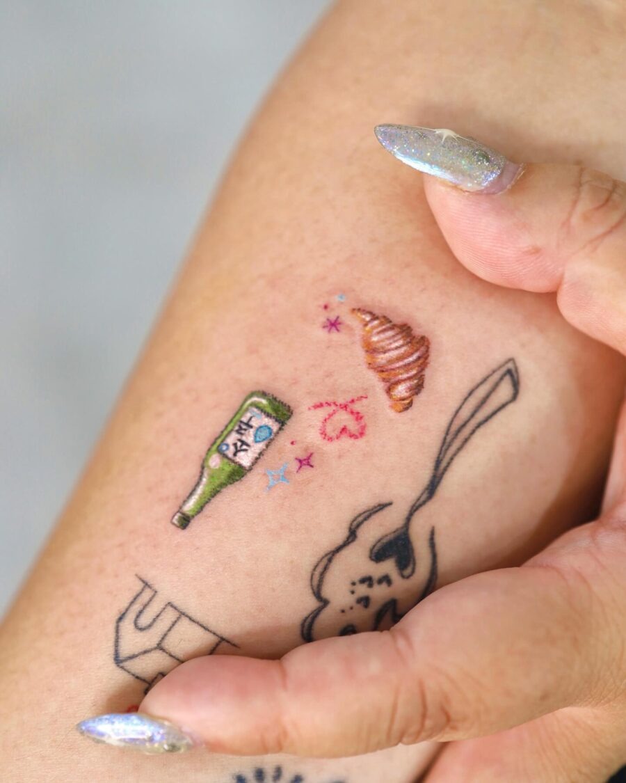 This charming tattoo combines a delicate croissant with a bottle and playful stars.