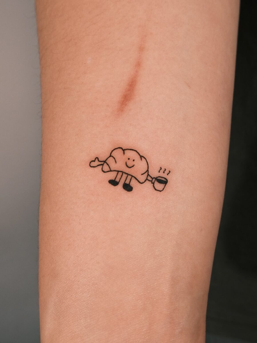 This adorable croissant tattoo features a smiling pastry character holding a steaming cup of coffee.