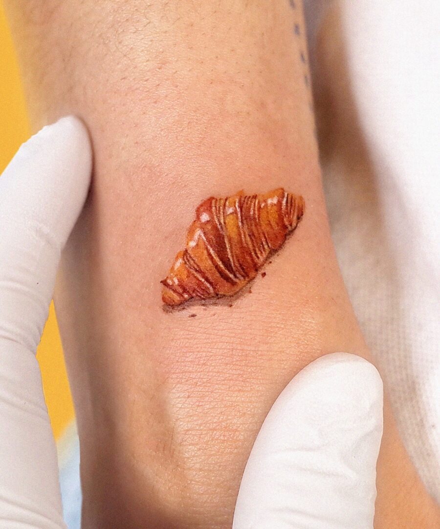 The tattoo showcases a beautifully realistic croissant, with intricate details capturing its flaky layers.