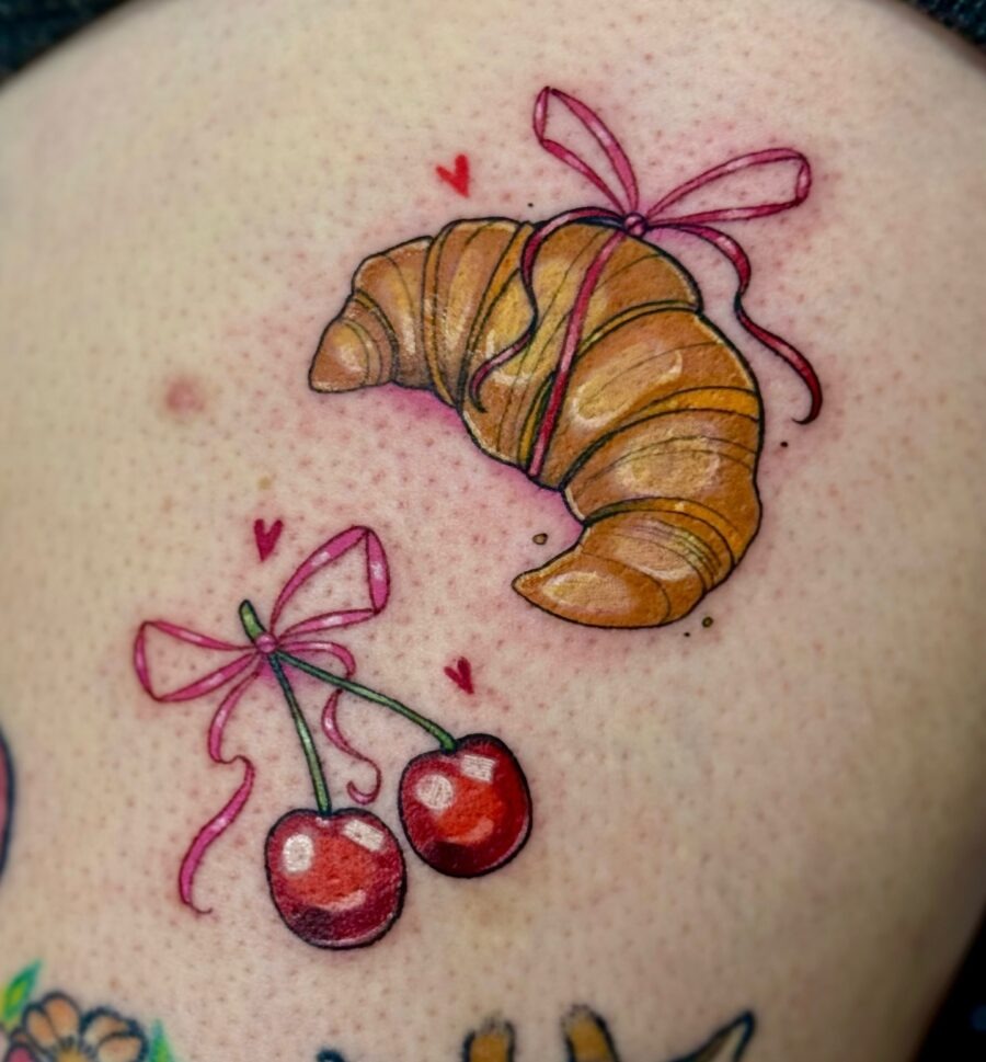This charming tattoo features a croissant adorned with a pink ribbon and matching cherry motifs.