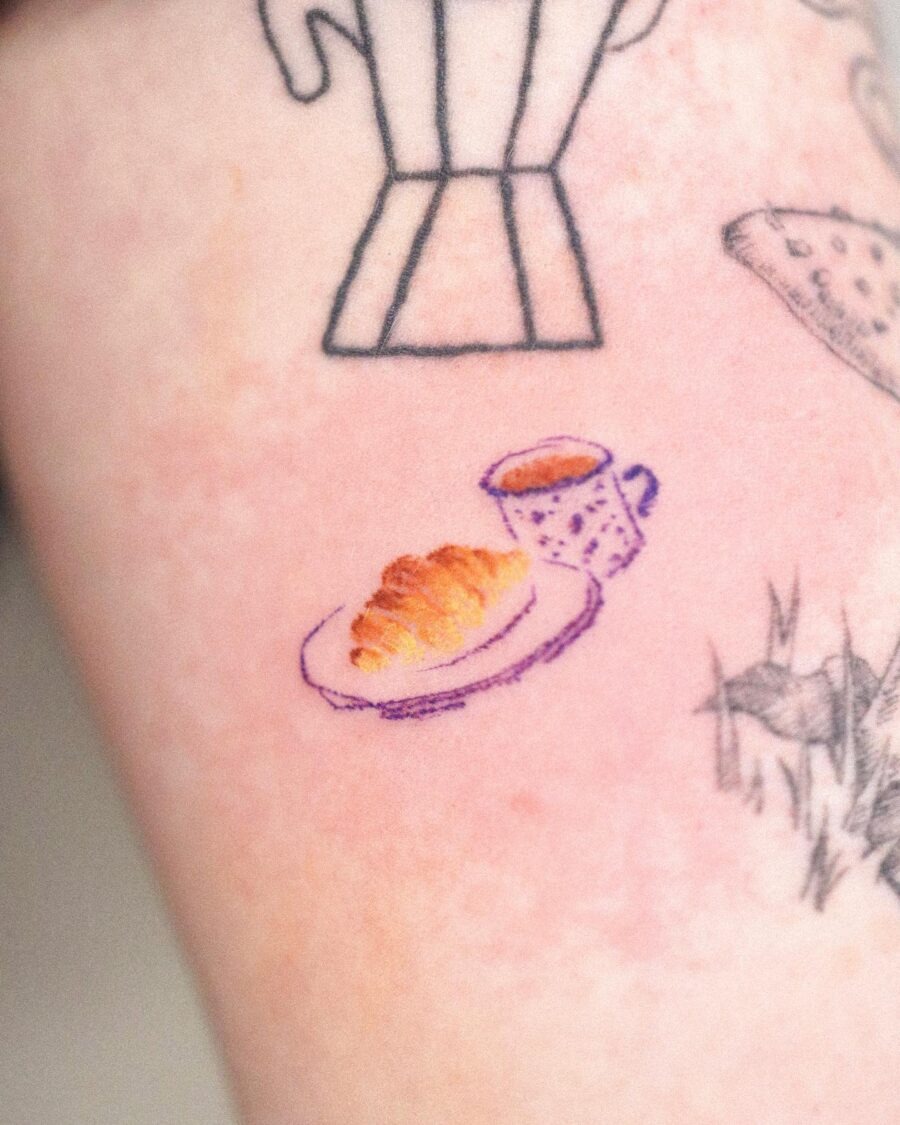 This charming tattoo showcases a golden croissant elegantly presented on a plate beside a cute coffee cup.
