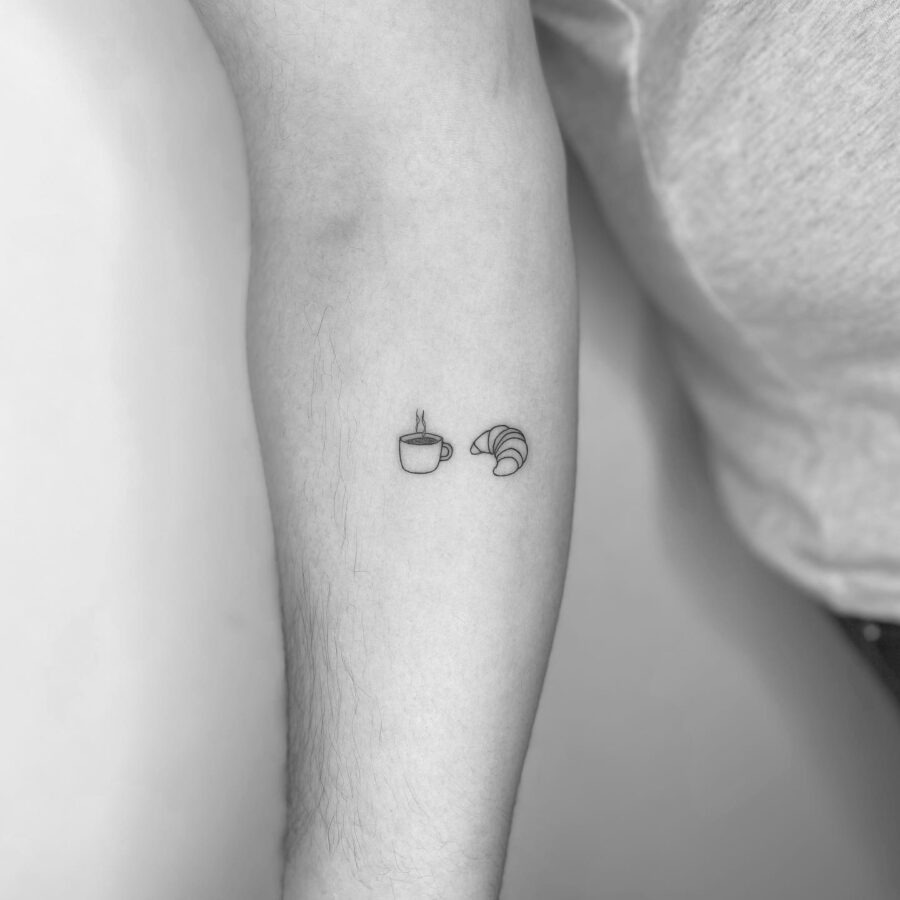 This minimalist tattoo features a delicate croissant alongside a coffee cup.