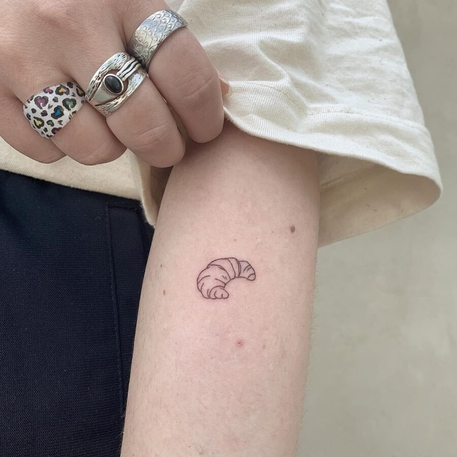 This charming tattoo features a simple outline of a croissant, nestled on the person's forearm.