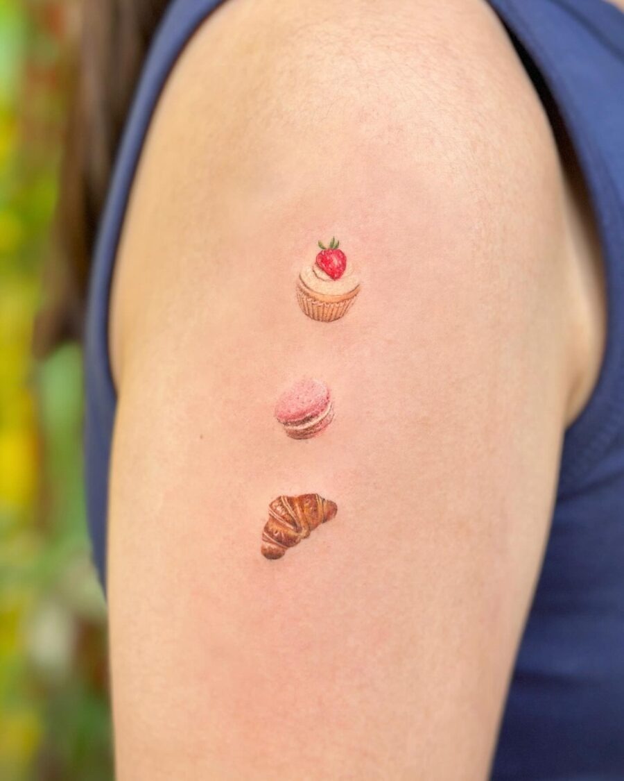 This tattoo showcases a trio of sweet treats, featuring a colorful cupcake, a pink macaron, and a beautifully detailed croissant.