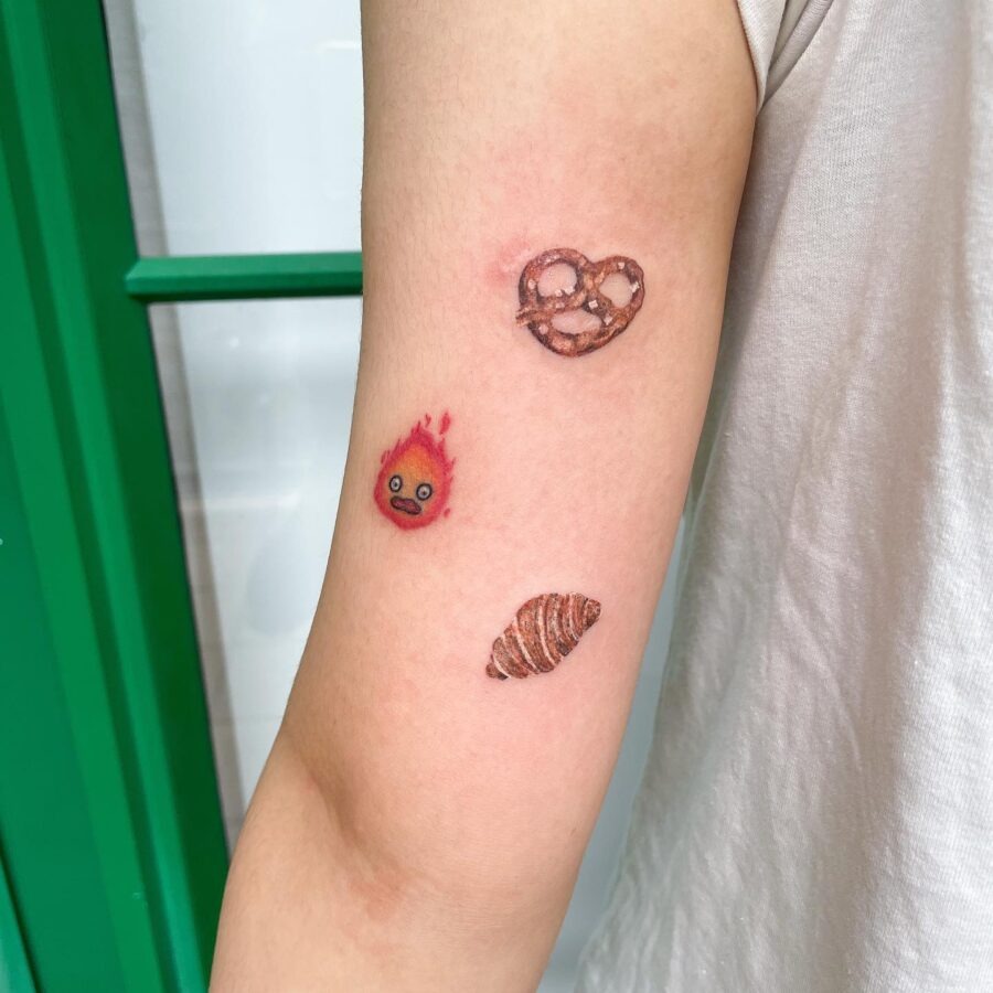 This tattoo design showcases a delightful croissant alongside a playful pretzel and an animated flame character.