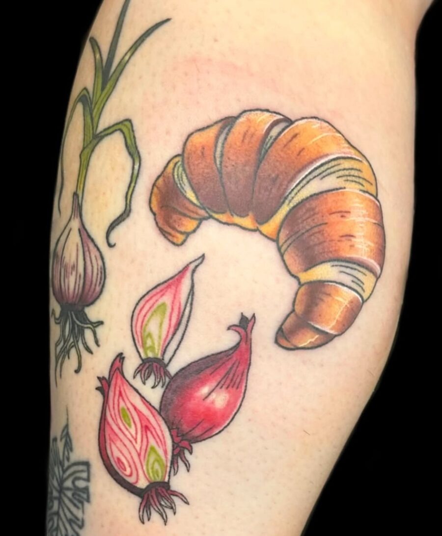 This tattoo features a detailed croissant alongside colorful onion bulbs.