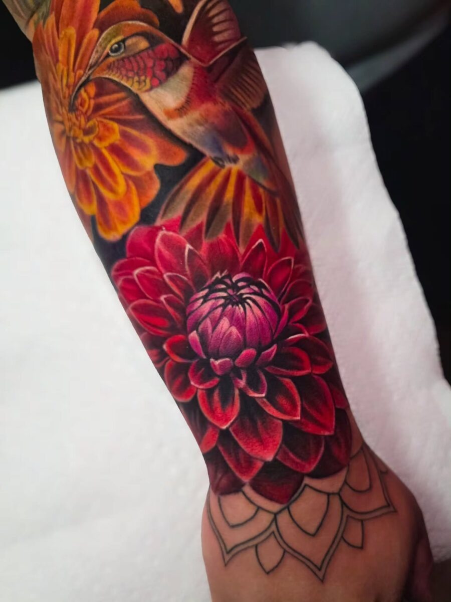 This striking tattoo features a bold dahlia in shades of red and pink, complemented by a beautifully detailed hummingbird.