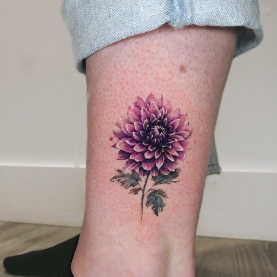 This stunning dahlia tattoo features purple hues, blending beautifully with the green leaves.