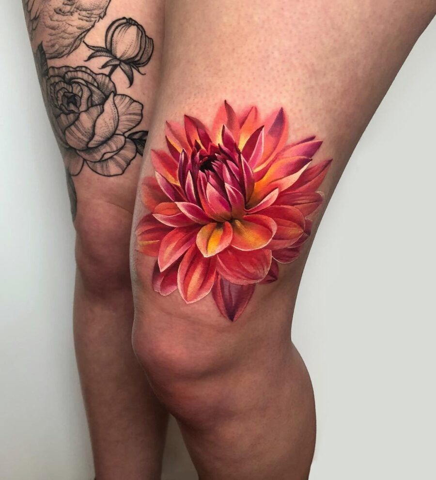 This dahlia tattoo features brilliant shades of orange and pink, creating a lifelike effect on the thigh.