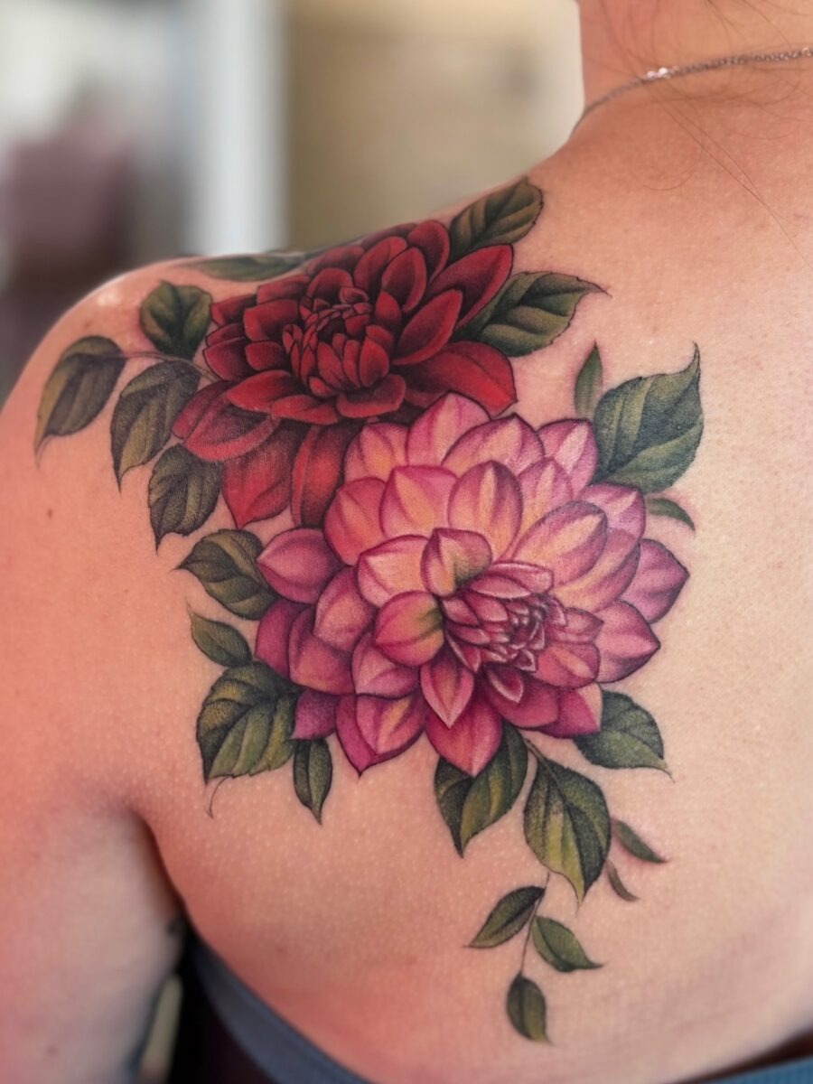 This tattoo showcases vividly colored dahlias with rich hues of red and pink, beautifully intertwined with lush green leaves.