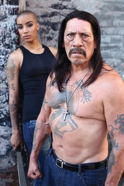 Trejo's intense gaze meets the camera, his chest tattoo on full display as he stands in front of a gritty, urban backdrop.
