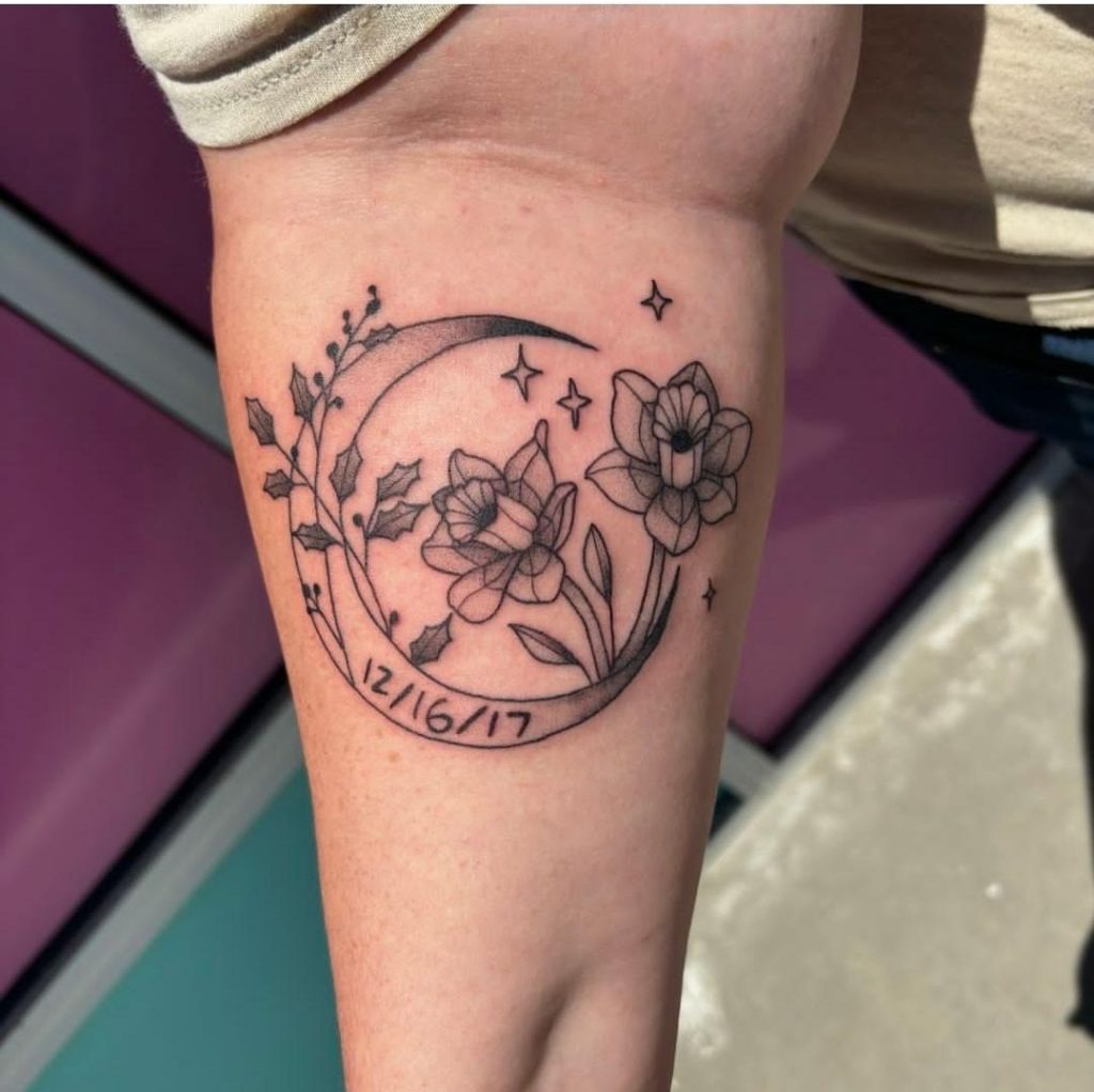This tattoo combines a crescent moon motif with Narcissus flowers and sprigs of holly, incorporating both December birth flowers into a celestial-inspired design.