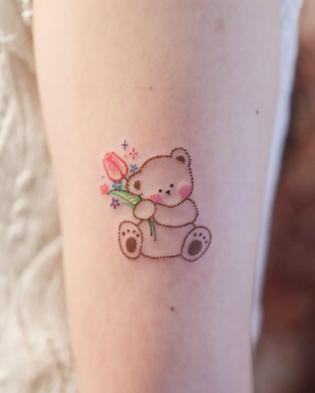 This charming bear tattoo showcases a cute bear gently holding a pink flower.