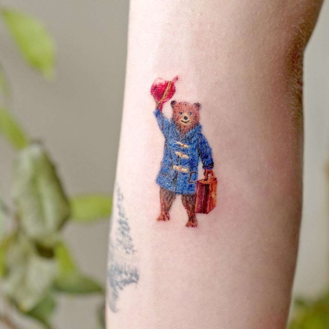 This tattoo features an adorable bear dressed in a blue coat, waving a red hat while holding a suitcase.