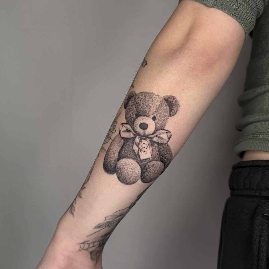 This captivating tattoo features a cuddly teddy bear adorned with a charming bow, perfectly inked on the forearm.