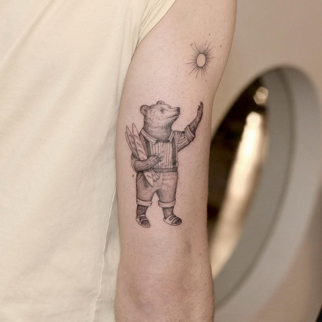 This tattoo features an adorable bear dressed in summer attire, holding a surfboard and waving at the sun.