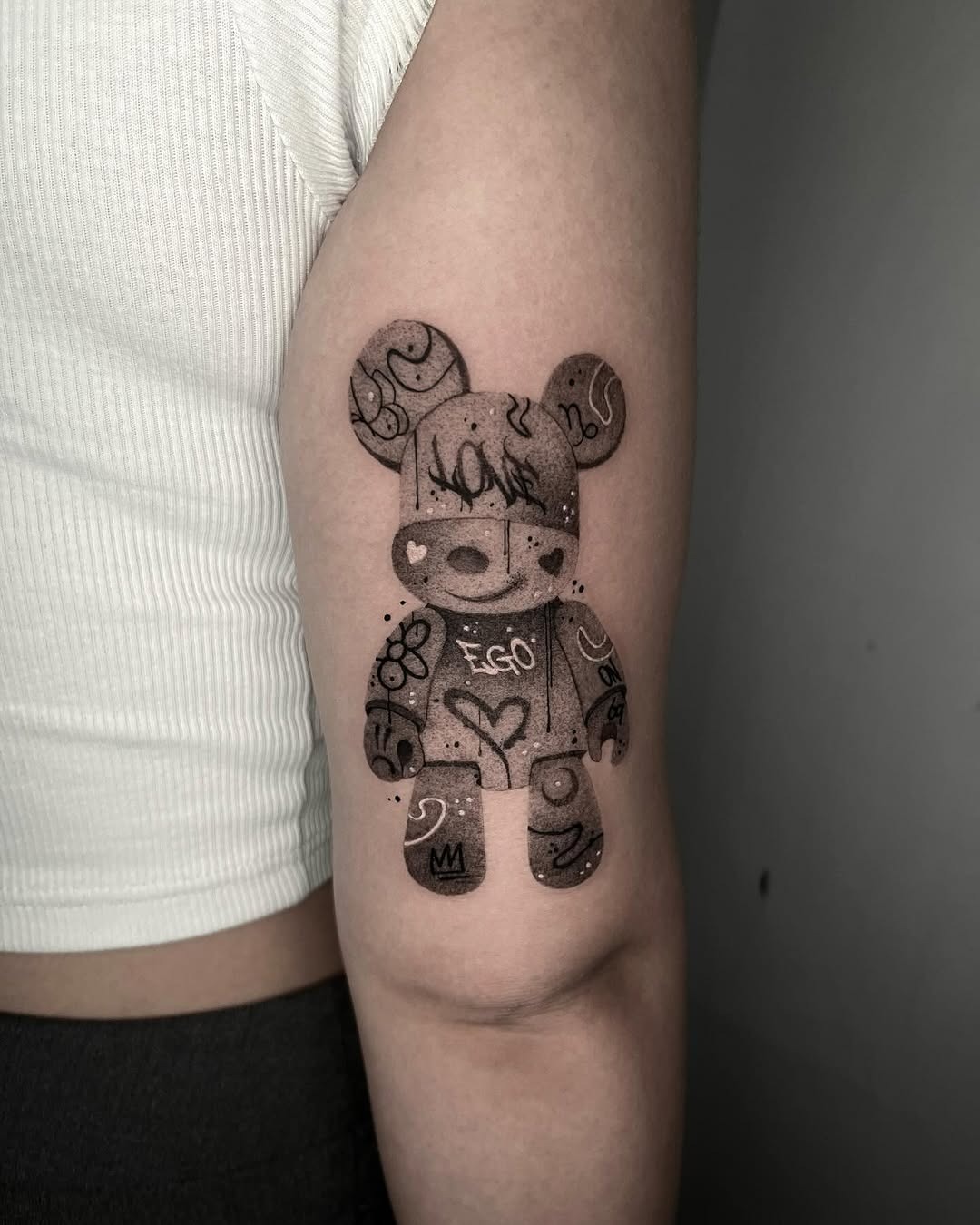 This tattoo depicts a cute bear character adorned with abstract designs and symbols, showing a blend of whimsical and artistic elements.