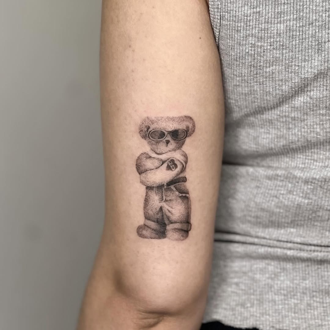 This bear tattoo features a cute, cartoonish bear wearing sunglasses and a relaxed pose.