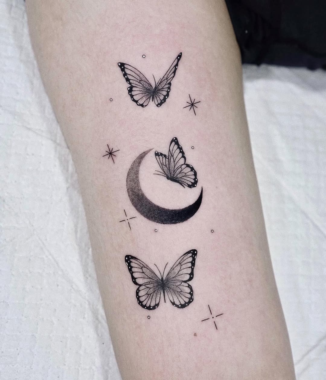 This tattoo features delicate butterflies fluttering around a crescent moon, interconnected by tiny stars.