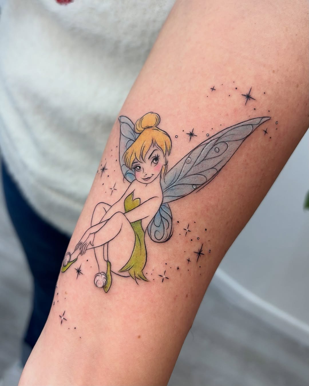 This tattoo showcases Tinkerbell perched playfully with her delicate wings.