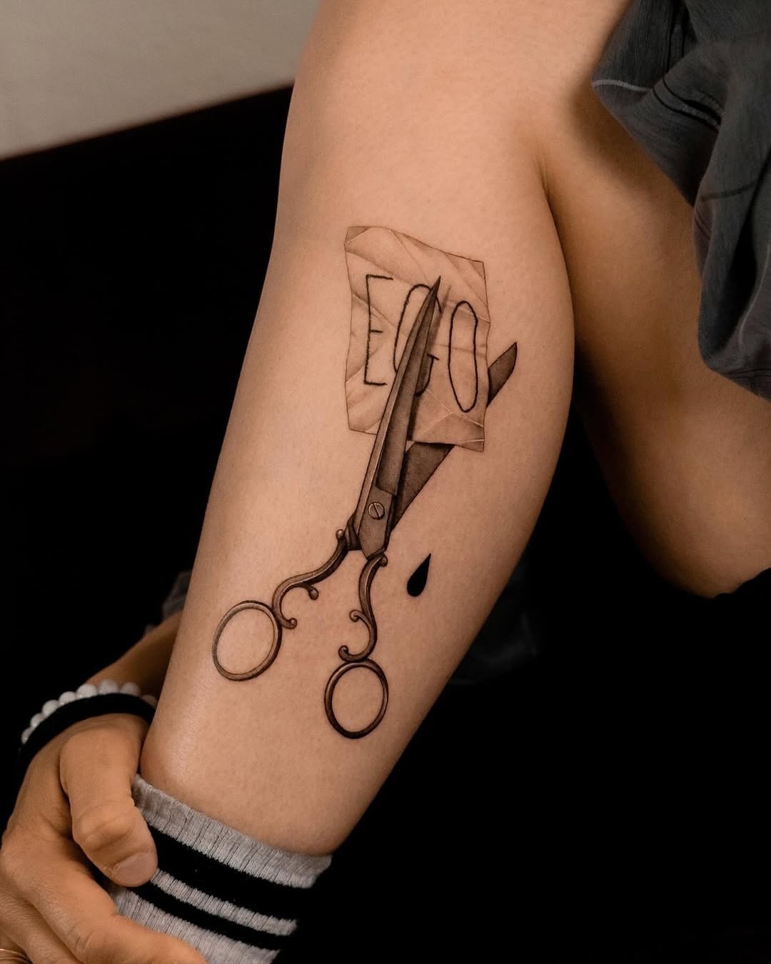 This unique tattoo features a pair of detailed scissors positioned above a sheet of paper.