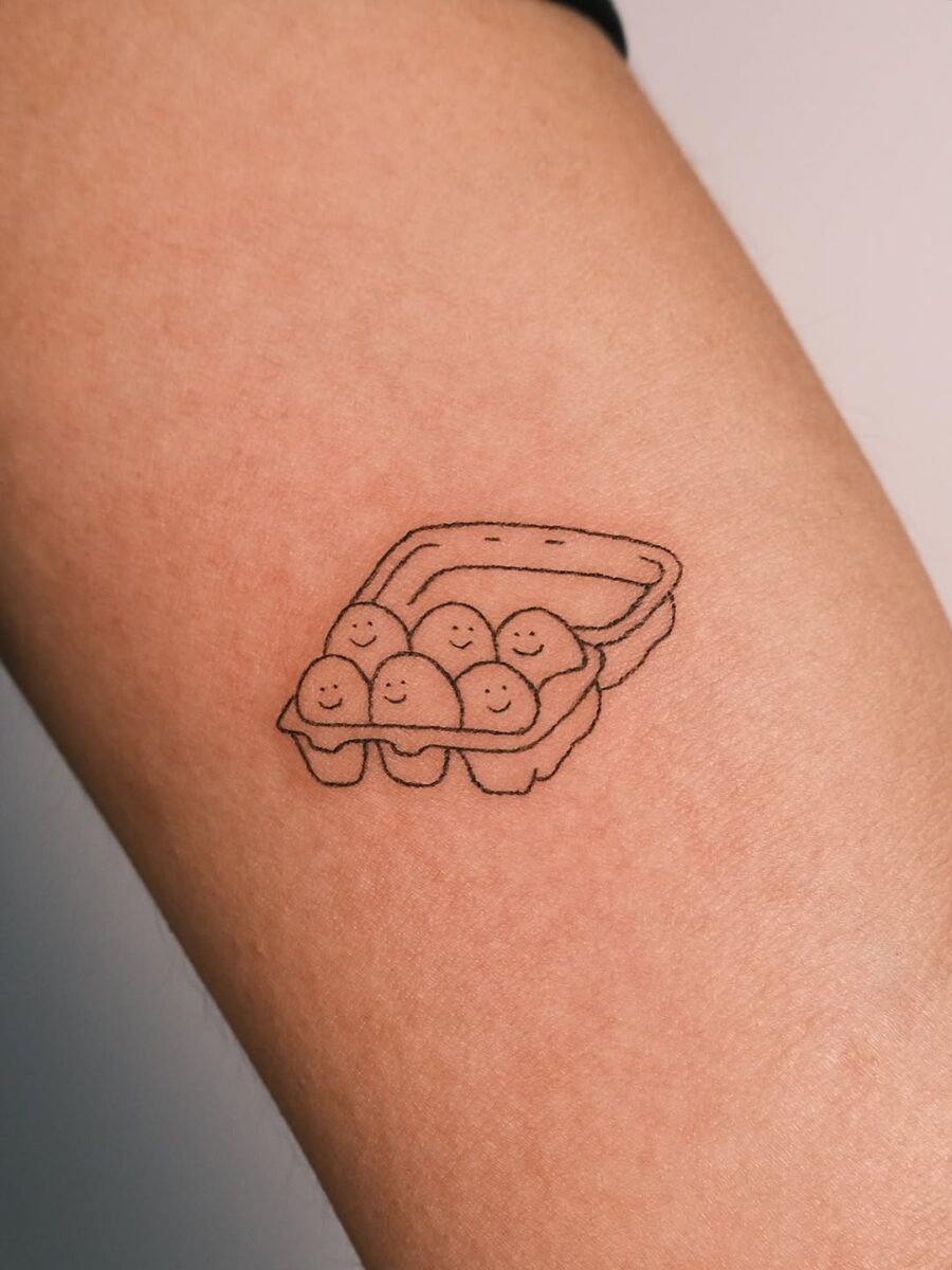 This whimsical tattoo features a cute egg carton, complete with smiling eggs peeking out.